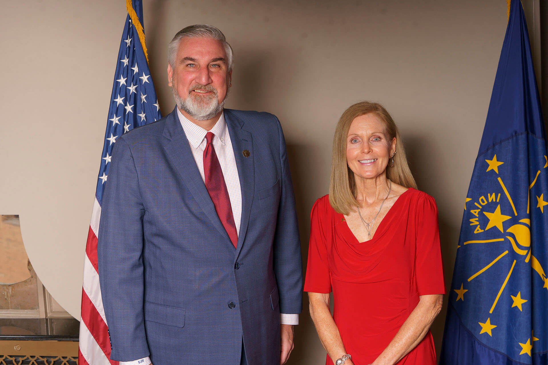 Lugar Series member wearing a red dress poses with Annual Luncheon speaker and flags