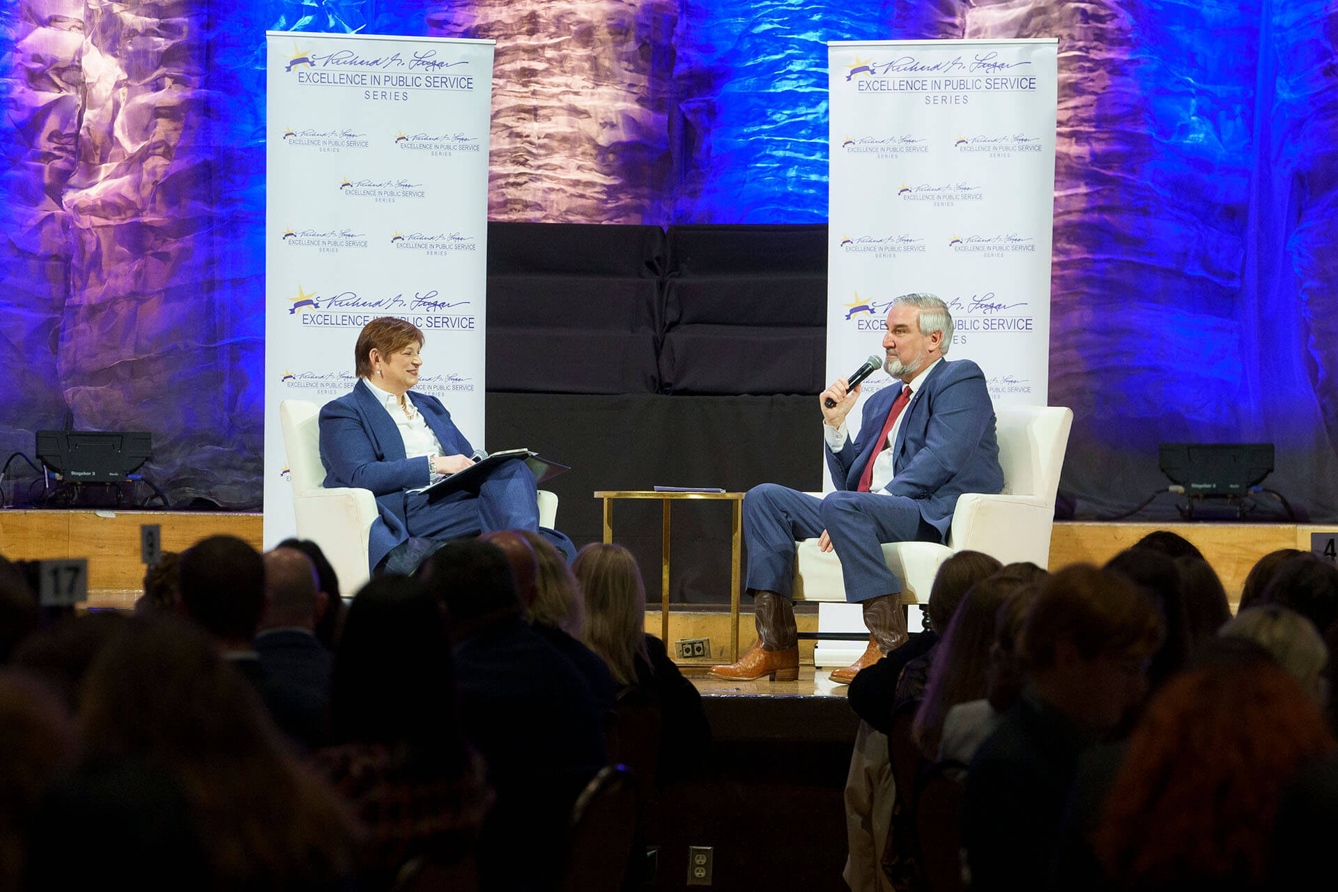 Lugar Series member and guest speaker have a conversation in front of Annual Luncheon attendees